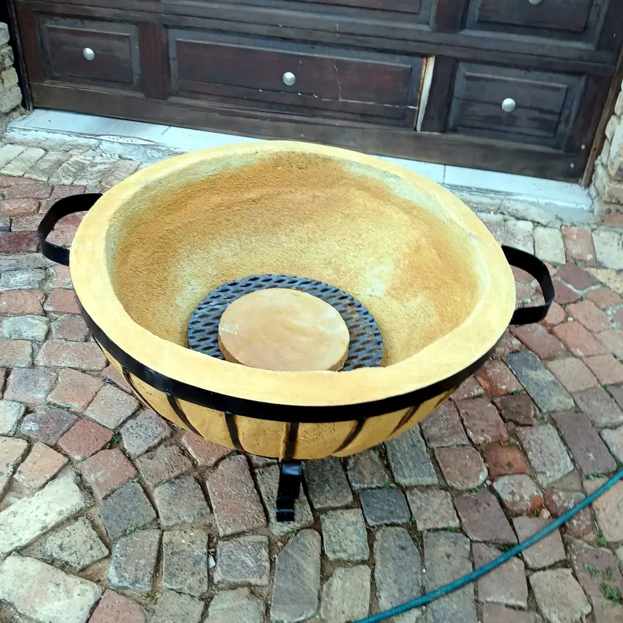 Close-up of an empty Motse boma braai bowl with its steel strap cradle, standing on a cobbled surface.