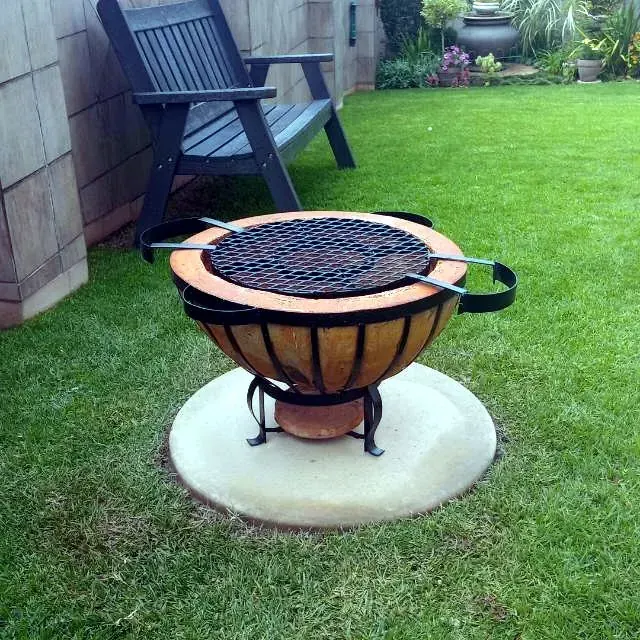 Motse boma braai fitted with a round cooking grid, set on a paved circle beside a lawn.